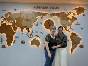 Deux femmes souriantes devant une carte du monde en bois avec horloge, représentant un voyage organi.