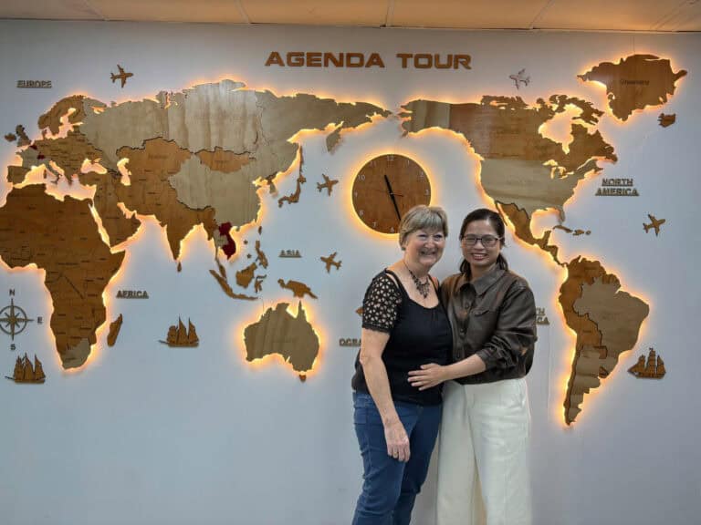 Deux femmes souriantes devant une carte du monde en bois avec horloge, représentant un voyage organi.